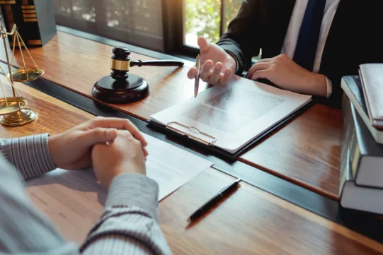 Two people sit at a wooden desk discussing legal documents. A gavel, scales of justice, and books are visible on the table.