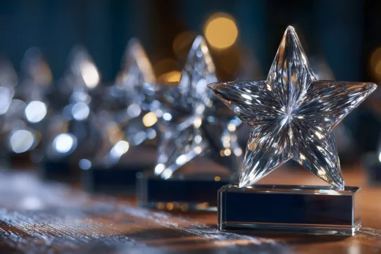 A row of clear glass star-shaped awards on a wooden surface with blurred lights in the background.