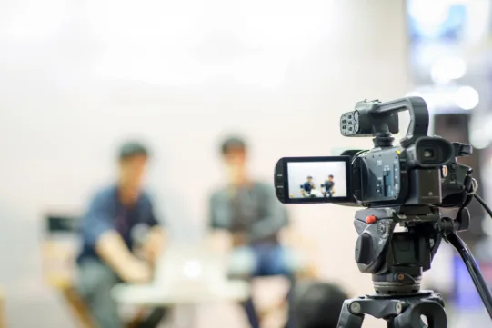 A video camera records two people sitting and talking in a blurred background, with the focus on the camera’s display screen.
