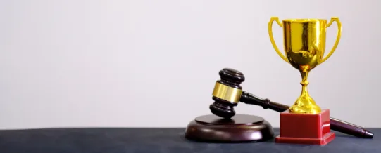 A wooden judge’s gavel rests against a gold trophy on a black surface, with a plain light background.