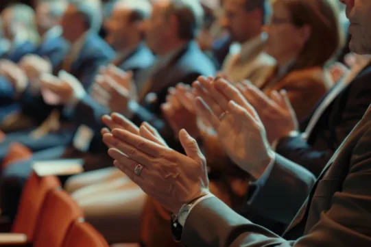 A group of people in business attire sit in rows, clapping their hands, suggesting applause at an event or conference.