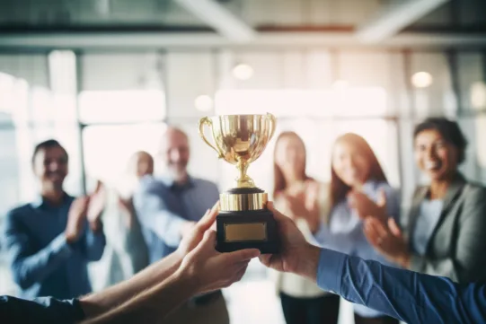 Two people hold a gold trophy while a group of people in the background clap and smile in an office setting.
