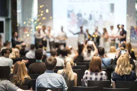 Audience seated, facing a stage where several people stand. Some attendees are taking photos. The background is blurred, highlighting the event’s formal or conference-like setting.