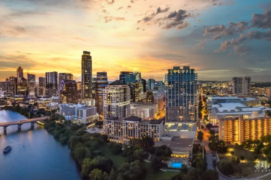Aerial view of a city skyline at sunset with tall buildings, a river, bridges, and clouds in the sky.