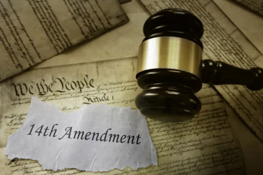 A judge’s gavel rests on documents with “14th Amendment” written on a torn paper; the U.S. Constitution is visible in the background.