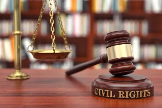 A wooden judge’s gavel labeled “Civil Rights” sits on a desk, with brass scales of justice and bookshelves blurred in the background.