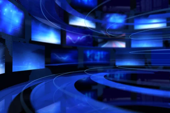 Futuristic digital studio with multiple blue-toned screens and curved reflective floor, resembling a modern television news set.