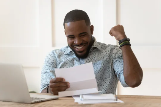 Smiling man sitting at a desk looks at a letter and raises his fist in excitement, with a laptop and papers in front of him.