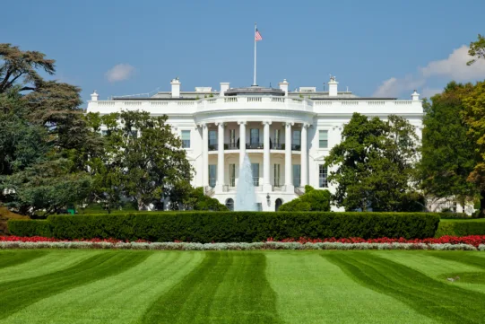 The White House with a green lawn and clear blue sky, viewed from the front with trees and flower beds in the foreground.
