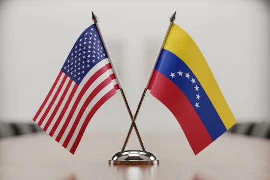 Two table flags, one of the United States and one of Venezuela, are crossed and displayed on a wooden table with a neutral background.