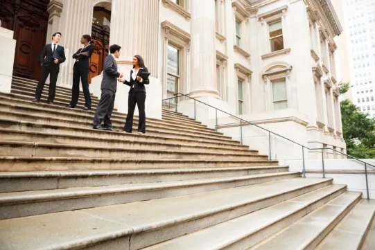 Four people in business attire stand and talk on the steps outside a large, classical building with columns.
