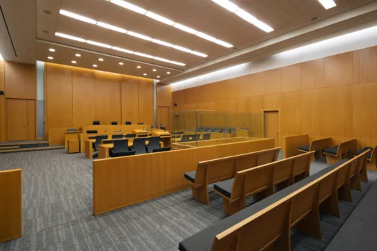 Empty modern courtroom with wooden benches, tables, judge’s bench, and glass enclosure under bright ceiling lights.