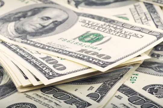 A pile of U.S. one hundred dollar bills, showing Benjamin Franklin’s portrait and various security features.