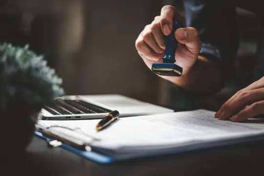 A person stamps a document on a desk next to a pen and an open laptop.