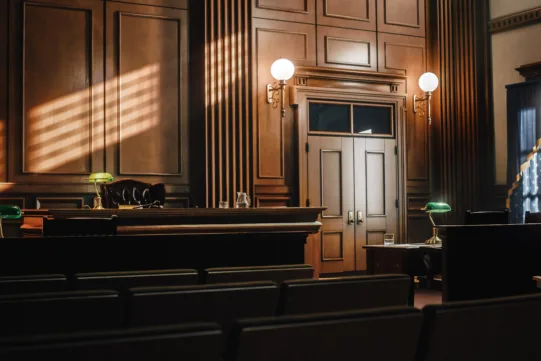 An empty courtroom with wooden walls, a judge’s bench, desks with green lamps, and sunlight streaming through the window blinds.