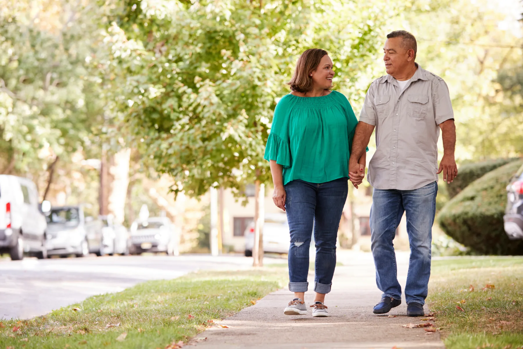 Home A middle-aged couple walks hand in hand on a sidewalk in a leafy suburban neighborhood, smiling at each other on a sunny day.