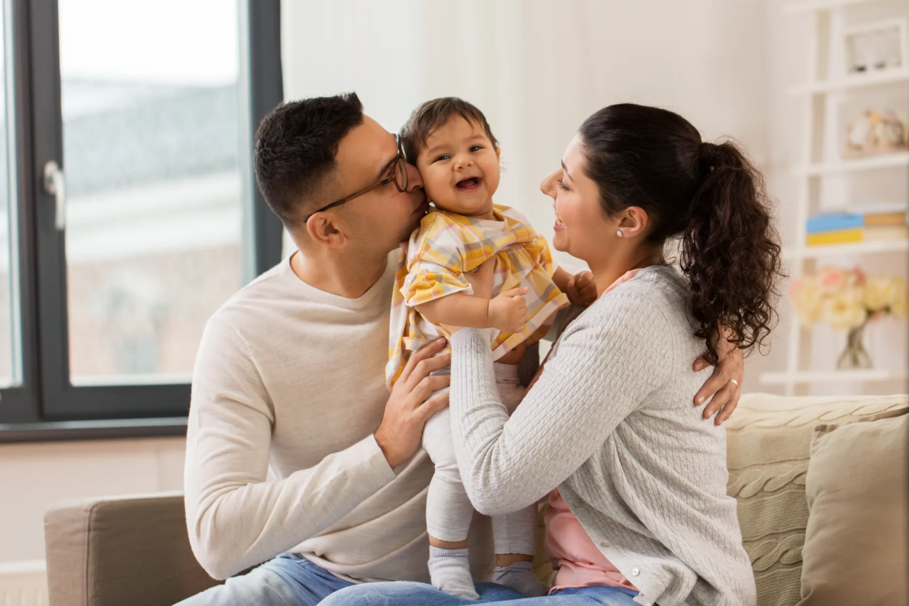 Home A man and woman sit on a couch, holding a smiling baby between them. The man kisses the baby’s cheek while the woman looks at the baby. A window and shelves are in the background.