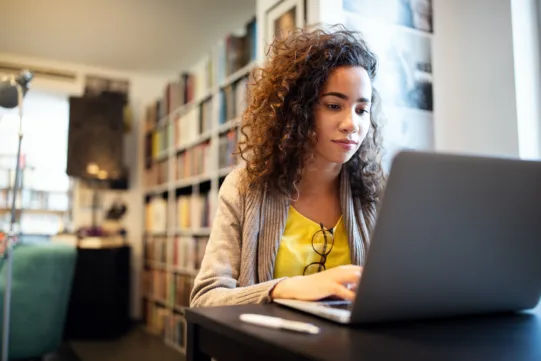 A student works on a laptop in a library, surrounded by bookshelves, while studying or conducting online research in a quiet learning environment.