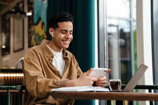 Young man wearing earphones smiles while looking at his smartphone at a table with a laptop, notebook, and glass of coffee in a well-lit cafe.