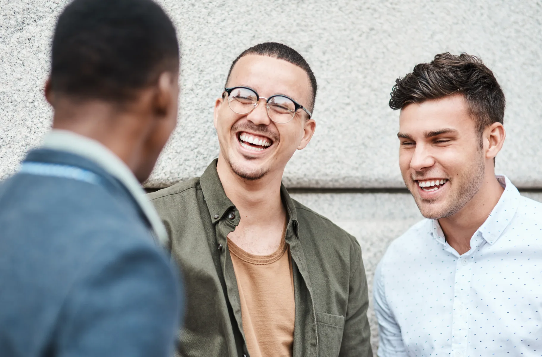 Home Three young men stand together outside, smiling and laughing as they have a conversation in front of a stone wall.