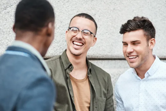 Three young men stand together outside, smiling and laughing as they have a conversation in front of a stone wall.