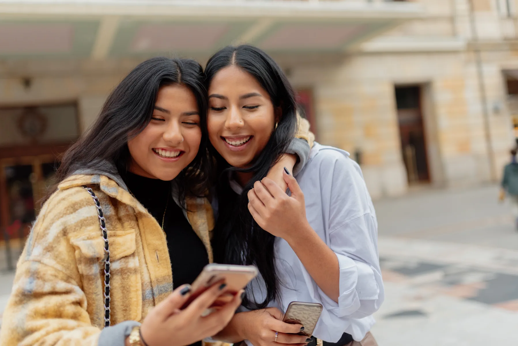 Home Two young women stand close together outdoors, smiling and looking at a smartphone, with buildings visible in the background.
