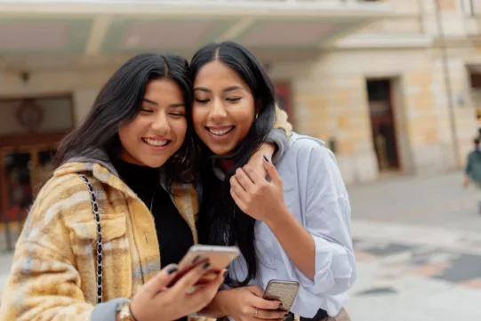 Two young women stand close together outdoors, smiling and looking at a smartphone, with buildings visible in the background.