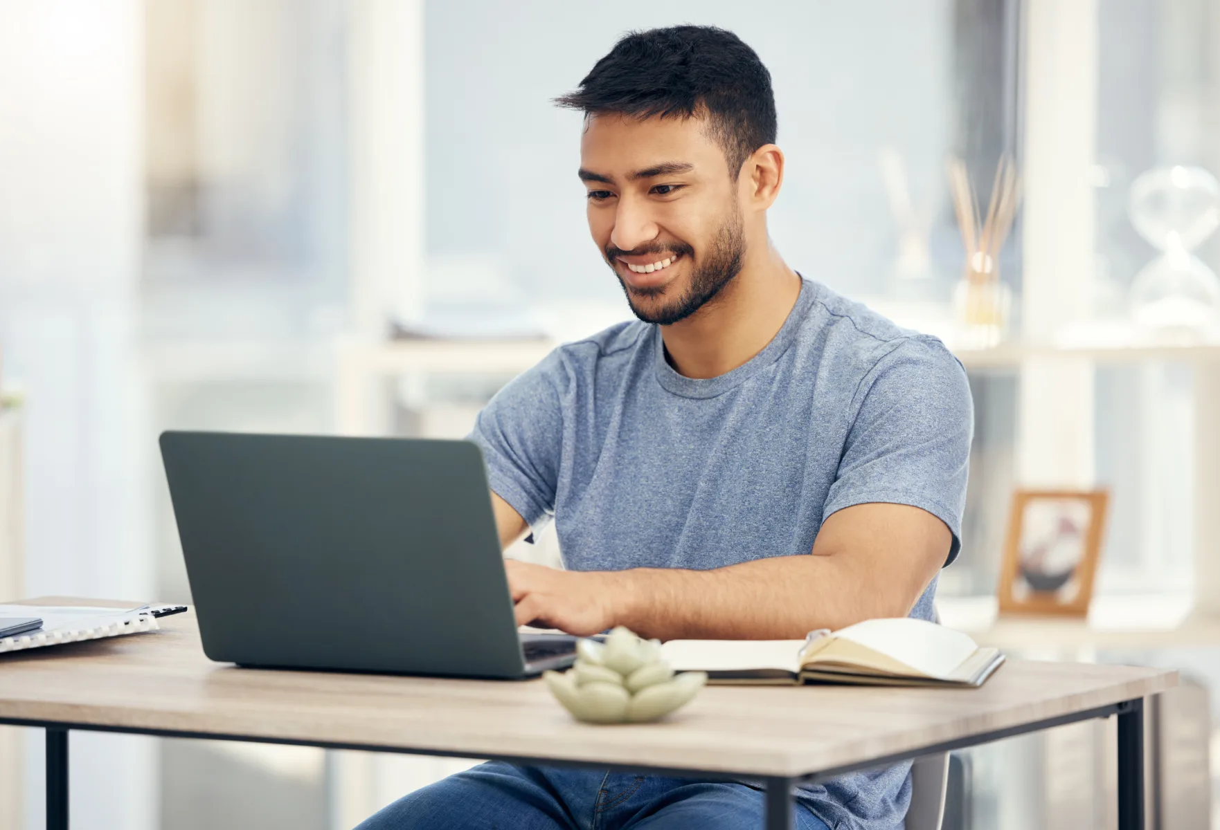 Home A man in a gray t-shirt sits at a desk, typing on a laptop, with an open notebook, pen, and a small plant nearby.