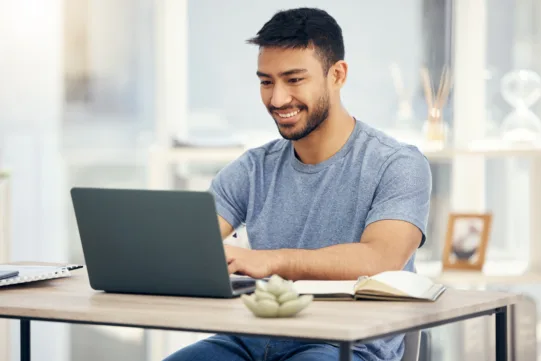 A man in a gray t-shirt sits at a desk, typing on a laptop, with an open notebook, pen, and a small plant nearby.