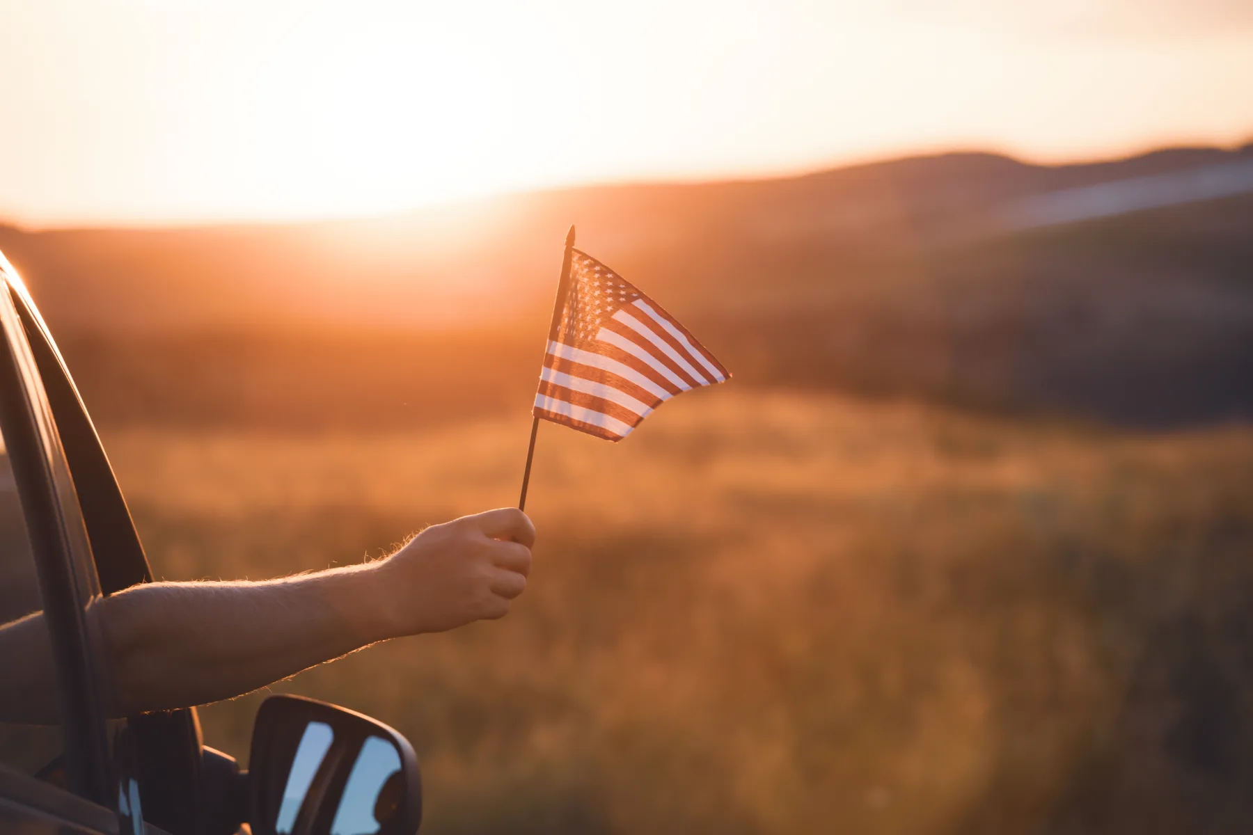 Home A person holds a small American flag out of a car window, with a grassy field and sunset in the background.