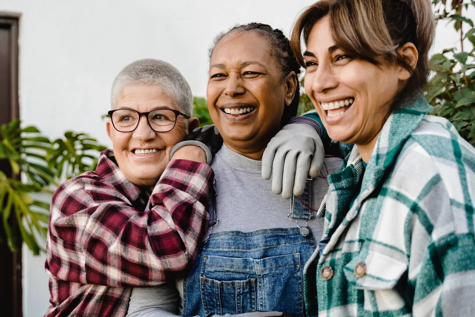 Home Three women in casual clothes stand close together outdoors, smiling and laughing. One woman wears gloves and overalls, another has glasses, and the third has brown hair tied back.