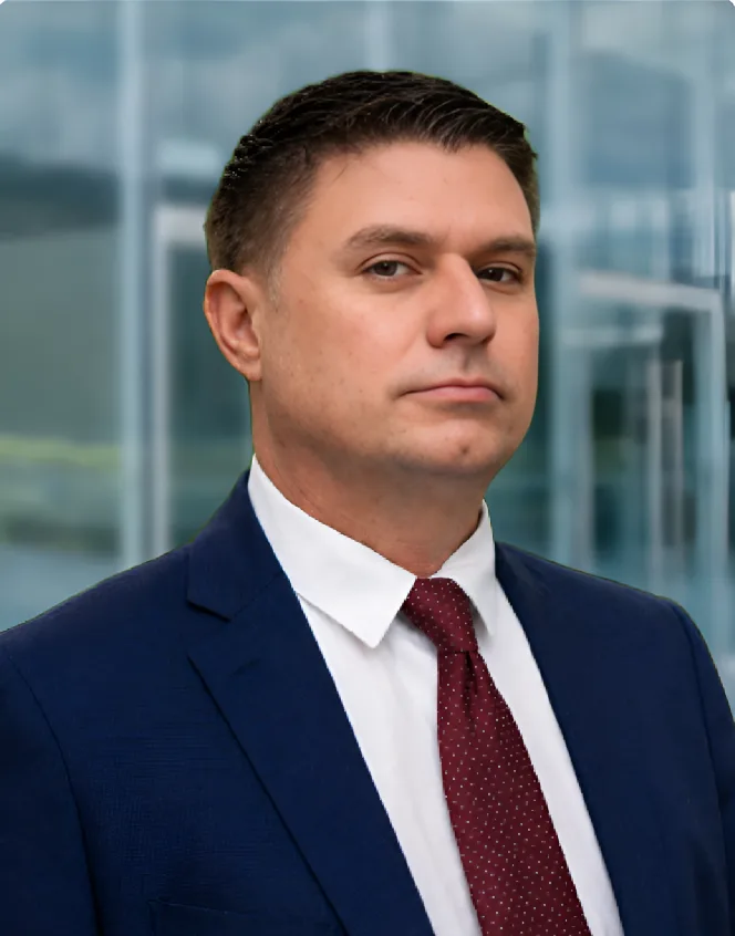 Marisol L. Pérez A man in a navy suit, white shirt, and red tie stands indoors in front of glass doors, looking at the camera with a neutral expression.