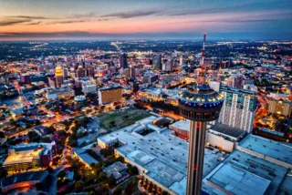 Home Aerial view of a city skyline at dusk featuring a prominent observation tower in the foreground and illuminated buildings in the background.