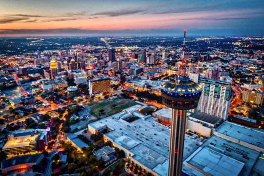 Aerial view of a city skyline at dusk featuring a prominent observation tower in the foreground and illuminated buildings in the background.