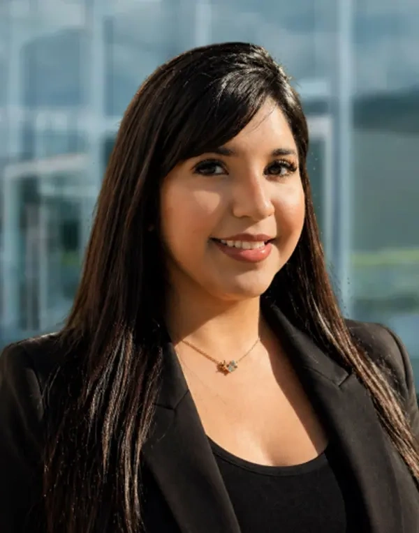 Marisol L. Pérez Woman with long dark hair wearing a black blazer and necklace, smiling and standing in front of a blurred glass building background.