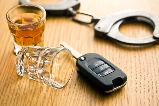 An amber-filled shot glass, an empty shot glass, a car key fob, and handcuffs on a wooden surface, relevant to a Houston deportation defense attorney.